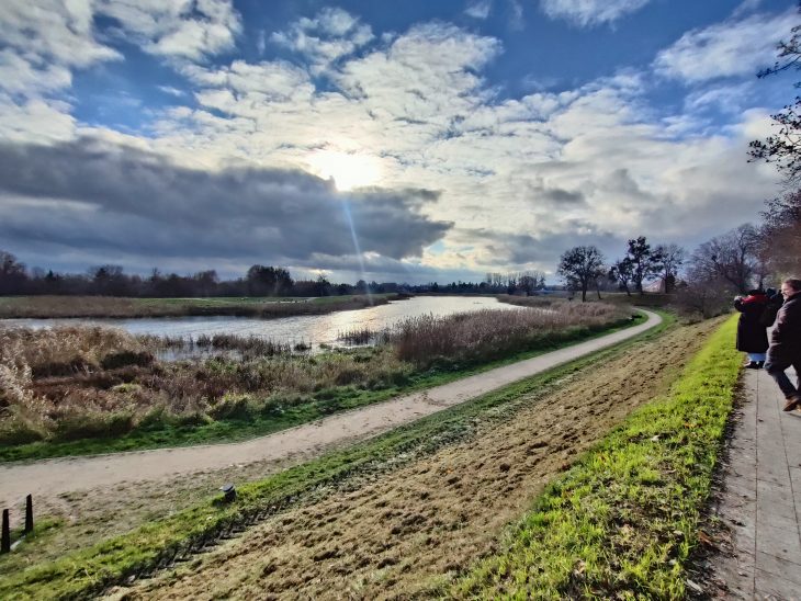 Przejście uczestników spaceru ze Śródmieścia do Dolnego Miasta w Gdańsku. Uczestnicy idą promenadą przy terenach zielonych, nadbrzeżem Opływu Motławy. Na zdjęciu widoczna część promenady zbudowana nie niewielkiej skarpie, poniżej której znajduje się ścieżka spacerowa oraz porośnięty wyższymi trawami brzeg Opływu Motławy. Niebo częściowo zachmurzone z widocznymi przebłyskami słońca.