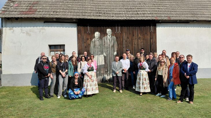 Grupa około trzydziestu osób stoi przed wiejską stodołą z dużym muralem przedstawiającym starszą parę. Dwie kobiety w grupie ubrane są w tradycyjne stroje ludowe z kwiecistymi spódnicami.