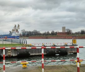 Na zdjęciu widoczne ujście Martwej Wisły odgrodzone barierkami od deptaka dla pieszych. Na rzece widoczny jest duży statek (masowiec) oraz mniejsze statki i łodzie. Po drugiej stronie rzeki, w tle, widoczna Twierdza Wisłoujście z charakterystyczną okrągłą wieżą z biało-czerwoną flagą na szczycie