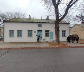 Uczestnicy spaceru oglądają odnowione budynki mieszkalne przy ul. Na Zaspę w Nowym Porcie w Gdańsku. Na zdjęciu widoczny dom z użytkowym poddaszem, szarą jednolitą elewacją oraz niebieskimi dwoma parami drzwi i ramami w oknach w tym samym kolorze. Na tle budynku, uśmiechnięty mężczyzna z rozpostartymi rękoma uśmiecha się do zdjęcia.