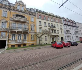 Zrewitalizowane kamienice przy ul. Rybołowców w Nowym Porcie w Gdańsku. Na pierwszym planie kamienica nr 8 z wyraźną żółtą ewaluacją i jaśniejszymi, piaskowymi zdobieniami przy okach i balkonach. Dalsze, również odnowione kamienice, są w kolorach bardziej stonowanych (zielonym i szary,) i mają mniej zdobień. Przed kamienicami widoczna droga z kostki oraz szyny z trakcją tramwajową.