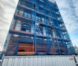 Odnawianie budynku w Śródmieściu w Gdańsku. Na zdjęciu widoczny odgrodzony stalowym ogrodzeniem wielopiętrowy budynek z ceglano-piaskową elewacją. Przy budynku stoi rusztowanie, a całość osłonięta jest siatką budowlaną.