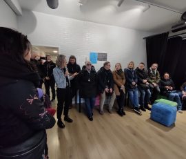 Uczestnicy spotkania w „Mrowisku” (budynku Towarzystwa Profilaktyki Środowiskowej) wsłuchujący się w wypowiedź przedstawicielki „Mrowiska”. Uczestnicy zgromadzeni w niedużej kwadratowej Sali z drewnianymi panelami na podłodze i białą cegiełką na ścianach. Część osób siedzi na kolorowych pufach, część stoi przy ścianach pomieszczenia.