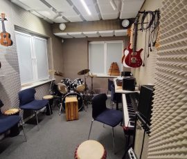 Pokój muzyczny w „Mrowisku” (budynku Towarzystwa Profilaktyki Środowiskowej), w którym dzieci objęte wsparciem mogą korzystać z instrumentów muzycznych. Na zdjęciu widać socjalnie przygotowane pomieszczenie – wygłuszone pianką ściany i sufit, dostępne instrumenty muzyczne: keyboard, bębny, gitary, perkusja oraz kilka krzeseł.