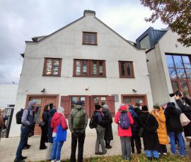 Uczestnicy spaceru zgromadzeni na chodniku przed domem sąsiedzkim (Inkubator Sąsiedzkiej Energii) przy ul. Reduta Wyskok w dzielnicy Dolne Miasto w Gdańsku. Budynek ma dwa pietra, zachowany tradycyjny kształt, odnowioną szarą elewację oraz drewniane, brązowe drzwi, ramy w oknach oraz okiennice przy oknach na dole budynku.