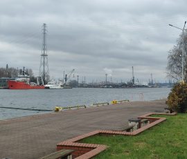 Na zdjęciu widoczna Martwa Wisła, na której zacumowano różnej wielkości statki. W tle, po drugiej stronie rzeki, widoczne są żurawie, koparki, wysokie słupy energetyczne, dymiący komin i inne zabudowania przemysłowe.