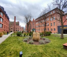 Zabudowania z czerwonej cegły lub z elewacją w kolorze ceglanym otaczająca i zielony wypielęgnowany plac przed ARCHE Dwór Uphagena w Dolnym Mieście w Gdańsku. Na alejkach placu widać kilka osób spacerujących, w tle wysoki wąski ceglany komin.