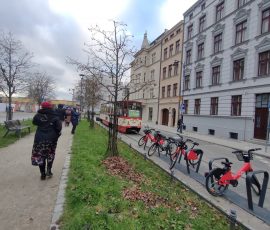 Spacer uczestników przez ścieżkę spacerową przy zbiegu ulic Wróbla i Toruńskiej w Gdańsku. Po prawej stronie widoczne są odnowione fasady kamienic. Na ulicy stoją miejskie rowery oraz nieczynny zabytkowy tramwaj z dużym banerem Opowiadacze Historii, który pełni funkcję zewnętrznej galerii dostępnej dla każdego.