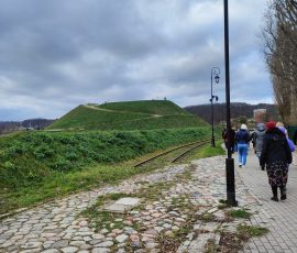 Spacer koło Bastionu Żubr w Gdańsku. Na zdjęciu widoczne zielone wzniesienie ze ścieżką i ławkami. Pomiędzy chodnikiem, po którym idą uczestnicy spaceru, a wzniesieniem (Bastionem Żubr) widoczne fragmenty torów tramwajowych.