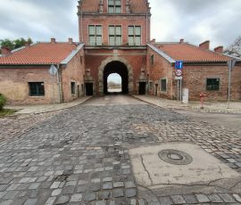 Brama Nizinna w Gdańsku. Budowla z czerwonej cegły. Dwa ramiona budynku stanowią niższe zabudowania ze skośnym dachem oraz w centralnej części brama z nadbudową dwupiętrową ze zdobieniami na najwyższym piętrze i na skraju dachu.