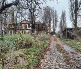 Jednopiętrowy budynek, który zostanie poddany rewitalizacji. Widoczne rozpoczęcie prac – zabudowano część wejść, okna zabezpieczono dyktami, rozmieszczono żółte tablice informacyjno-ostrzegawcze. Budynek znajduje się w zarośniętym krzewami i drzewami ogrodzie.