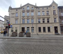 Zrewitalizowana elewacja w kolorze jasno-żółtym z zachowanymi zdobieniami przy oknach. Odnowiony budynek kontrastuje ze starym, szarym i nieodnowionym budynkiem, który częściowo widoczny jest na zdjęciu po prawej stronie. Przed budynkiem widoczny chodnik, przejście dla pieszych z sygnalizacją świetlną, przechodnie i ulica wyłożona szarą kostką kamienną