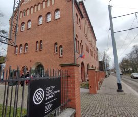 Piętrowy budynek z czerwonej cegły, w którym mieści się Centrum Sztuki Współczesnej Łaźnia 2, o czym informuje czarna tablica z białymi napisami na stalowym ogrodzeniu