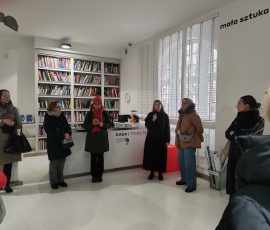 6. Uczestnicy spaceru po obszarze rewitalizacji w Centrum Sztuki Współczesnej Łaźnia 2 przy kasie biletowej, słuchający pracownicy Centrum, która mówi przez niewielki mikrofon. W tle, za kasą, widoczny duży regał z książkami