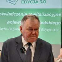 Portret starszego mężczyzny w ciemnym garniturze i krawacie, stojącego na tle ścianki z napisem „Regiony Rewitalizacji Edycja 3.0”. Do klapy marynarki przypięty ma niewielki mikrofon bezprzewodowy.